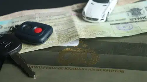 Close-up of a car key, a remote control, a white toy car, and Indonesian vehicle registration documents, symbolizing the smooth process of Car Ownership Transfer.