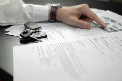 A person points to documents spread on a table alongside car keys, preparing for the vehicle title transfer.