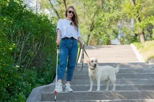 A woman in sunglasses and casual clothing stands on stairs with a guide dog. She holds a white cane in one hand and the dog's leash in the other. The background shows greenery and trees, reflecting the beautiful landscapes of Australia.