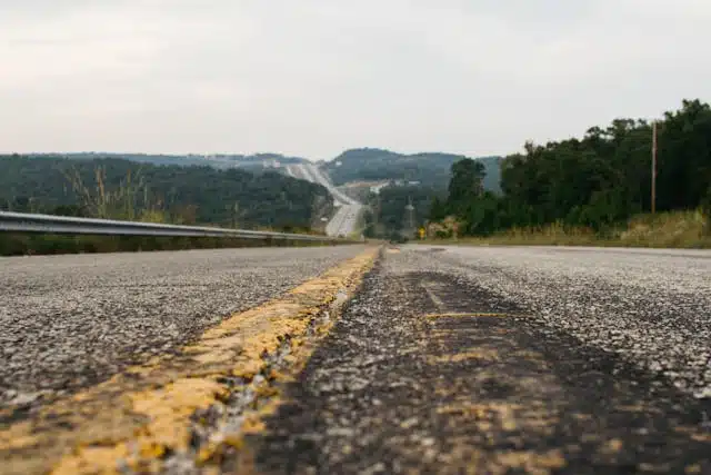 A low-angle view of a rural road with a worn yellow line down the center evokes the freedom of a holiday road trip, leading towards distant hills and surrounded by trees under an overcast sky.
