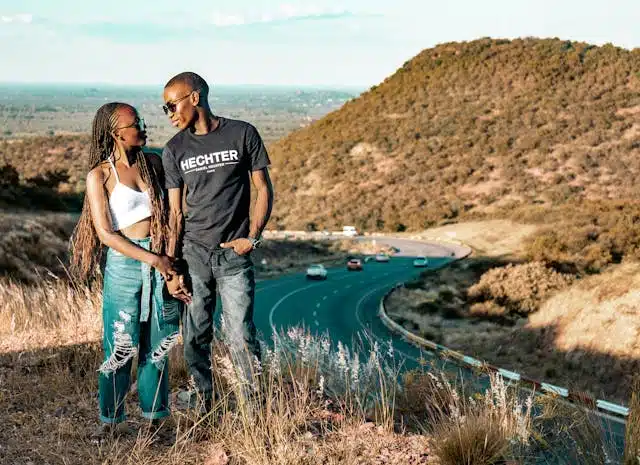 A couple stands on a grassy hill overlooking a winding road with cars.