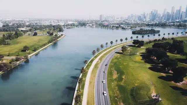 An aerial view captures a serene lake with a curving road perfect for a holiday road trip, bordered by lush green lawns and trees. The city skyline stands in the distance under a clear sky, offering breathtaking views along your journey while ensuring road trip safety.