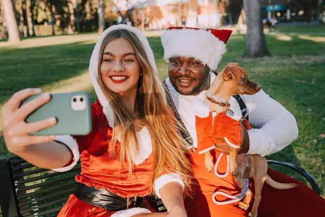 Two people in Santa outfits take a selfie on a bench in a park, with one holding a small dog dressed in red, all ready for the Christmas dinner festivities.