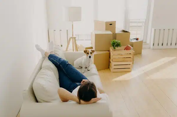 A person relaxes on a white couch, their small dog by their side, embodying the joys of being pet parents. Around them, cardboard boxes and a wooden crate are stacked in the brightly lit room, hinting at a move amidst changing rental laws.