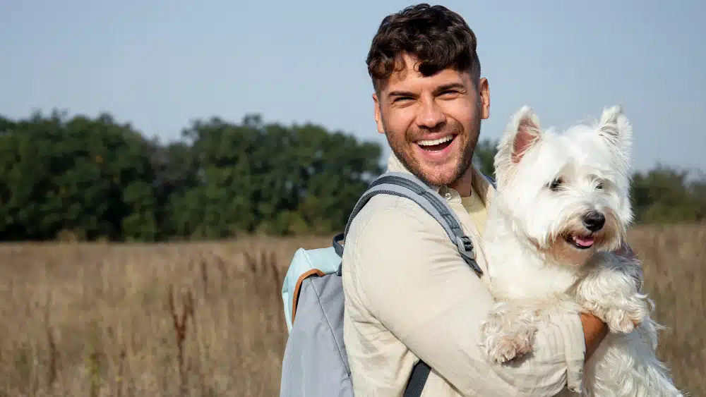 A man with a backpack smiles while holding a white dog in his arms in an open field with trees in the background, perhaps reflecting on pet plans or how to choose pet insurance for his loyal companion.