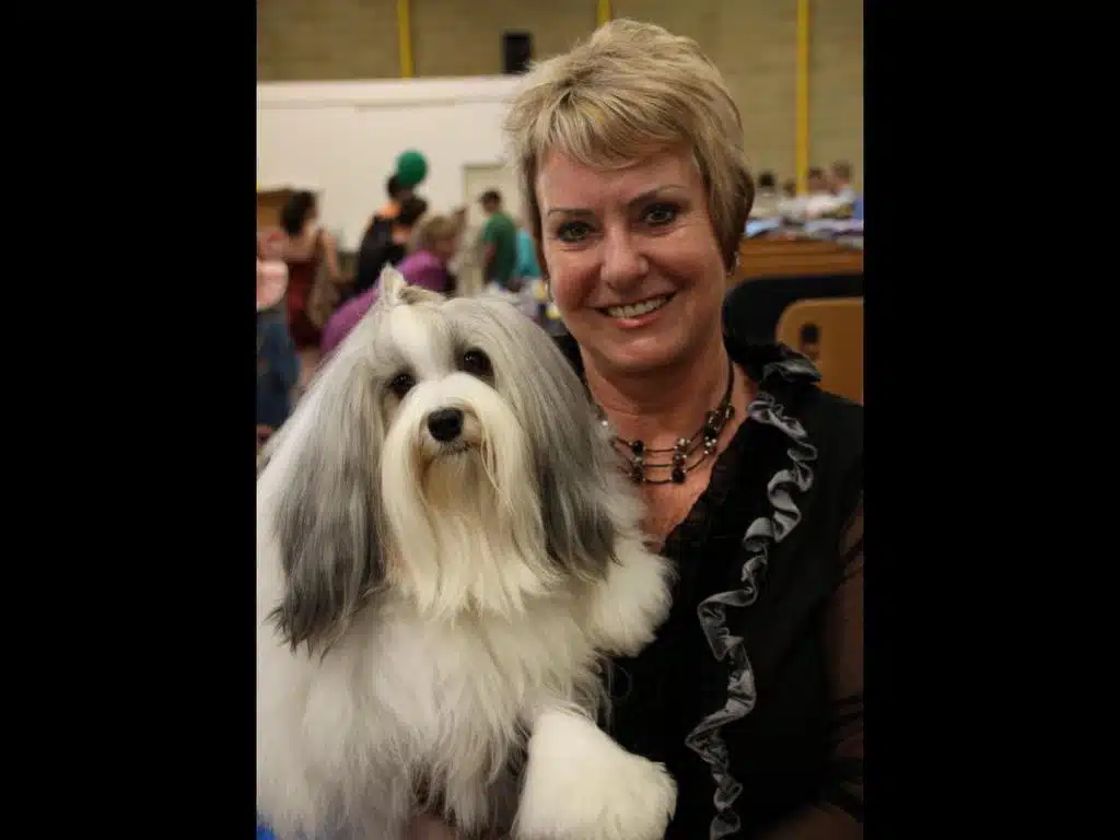 At a dog show, a woman cradles her fluffy white and gray companion, with blurred attendees mingling in the background.