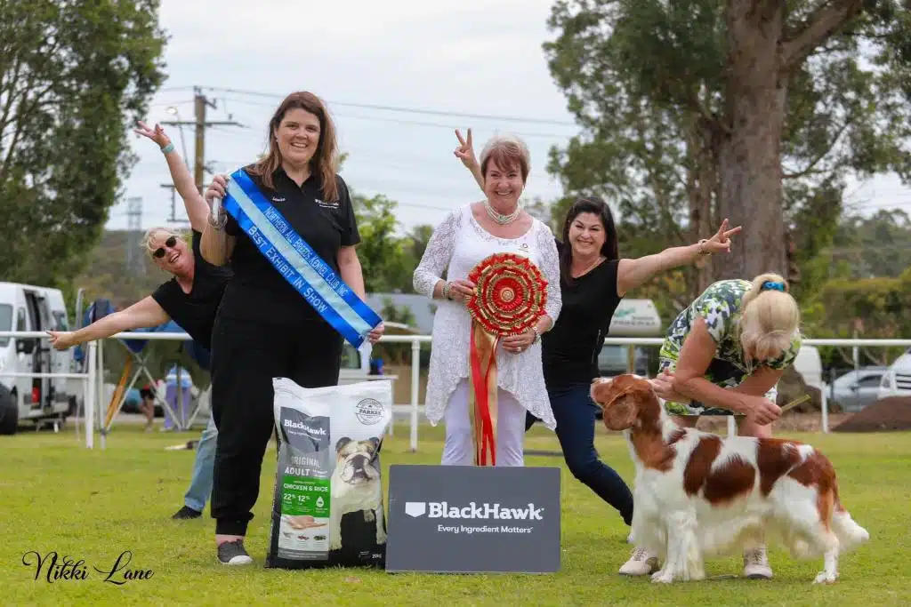 A group of people celebrates joyfully with a dog, ribbons, and dog food in front of a Black Hawk sign at an outdoor dog show event.