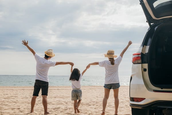 A family of three stands on a beach, facing the ocean, with the adults wearing hats and holding their child's hands. An open car trunk is visible on the right—a reminder that Easter road safety starts with mindful driving and secure packing.