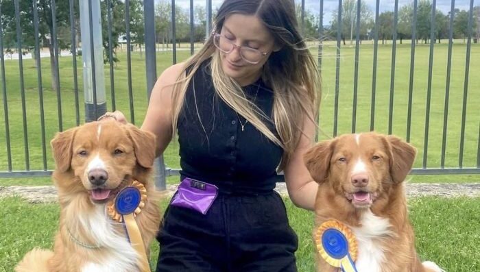 Dogs West member, Jess, kneels on grass with her two duck toller pups, who are both involved in dog sports