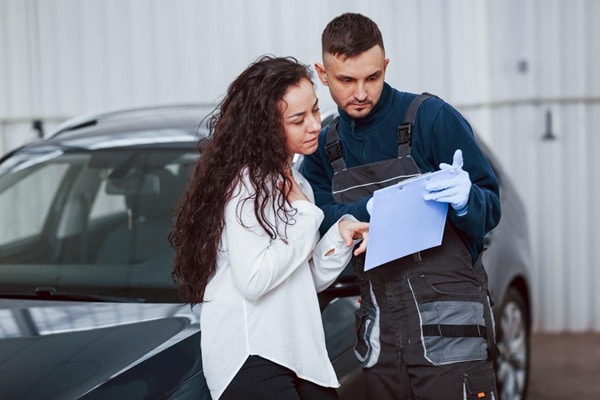 A mechanic in overalls and gloves shows a clipboard to a woman standing next to a car inside a garage, discussing how often should you service a car for optimal performance and safety.