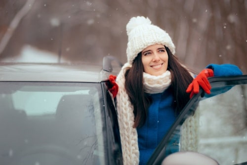 A woman wearing a white hat, scarf, blue coat, and red gloves smiles while standing by an open car door in a snowy outdoor setting, ready to save petrol and reduce car costs this winter.