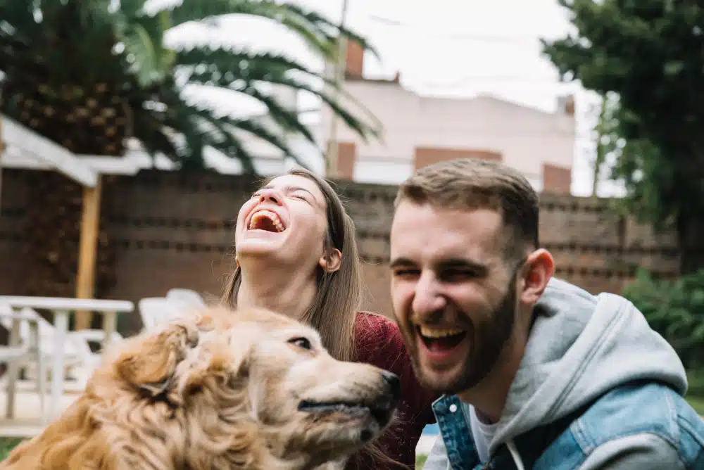 A man and woman are laughing while sitting outdoors with a golden retriever in the foreground, celebrating International Dog Day. Trees and patio furniture are visible in the background.