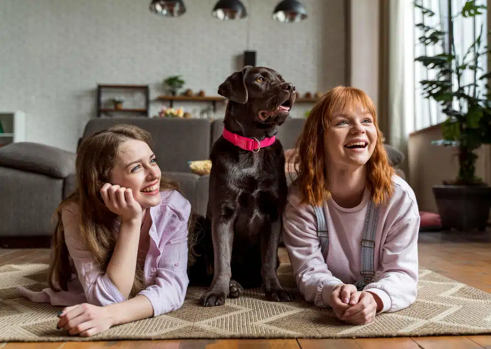 Two young women lie on a rug indoors next to a chocolate Labrador with a pink collar; all three are looking forward and smiling, celebrating International Dog Day together.