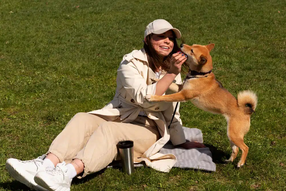 A woman sits on the grass with a tumbler, smiling as a Shiba Inu stands on its hind legs.