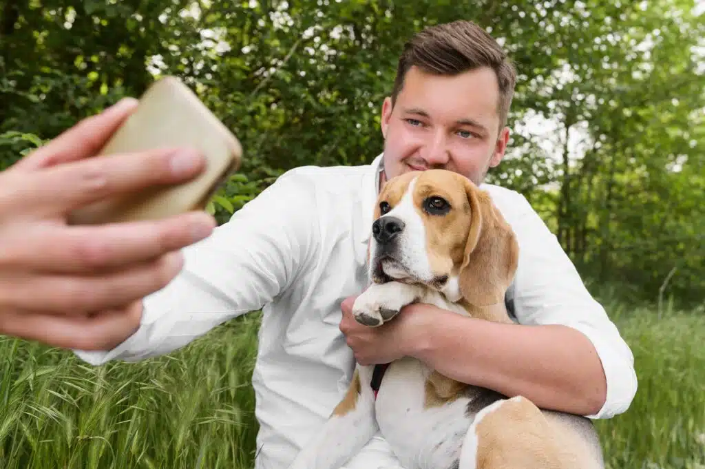 A man in a white shirt sits on grass, holding a beagle dog and taking a selfie with a smartphone outdoors, celebrating International Dog Day.